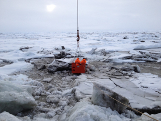 This image shows sediment-rich sea ice in the Transpolar Drift Stream. A crane lowers two researchers from the decks of the icebreaker RV Polarstern to the surface of the ice to collect samples. Photo Credit: R. Stein, Alfred Wegener Institute