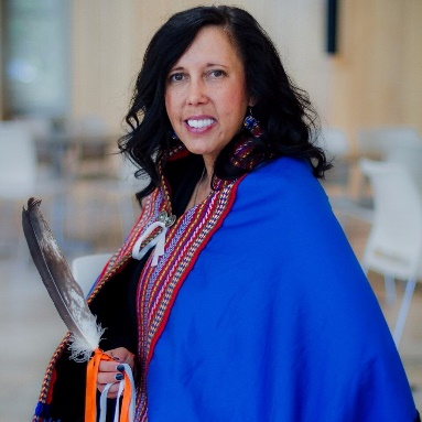 Dr. Carrie Bourassa: a middle-aged woman with wavy dark hair wearing traditional regalia (beaded earings, a blue cape with embroidery) and holding a feather